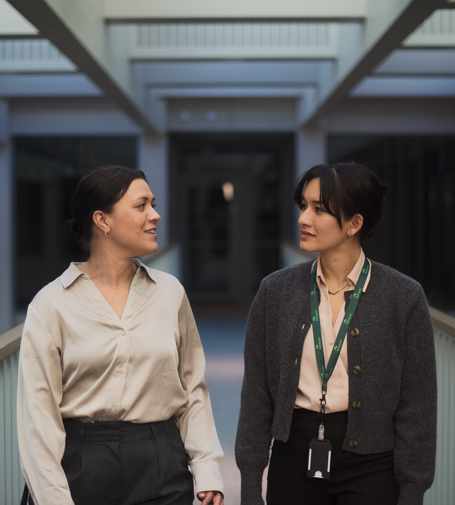 Two people walking side by side indoors, talking, with a modern office corridor in the background.