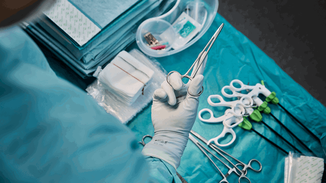 The hand of a surgeon holding a clamp over other surgical tools and draping laid out on a table. Photo. 