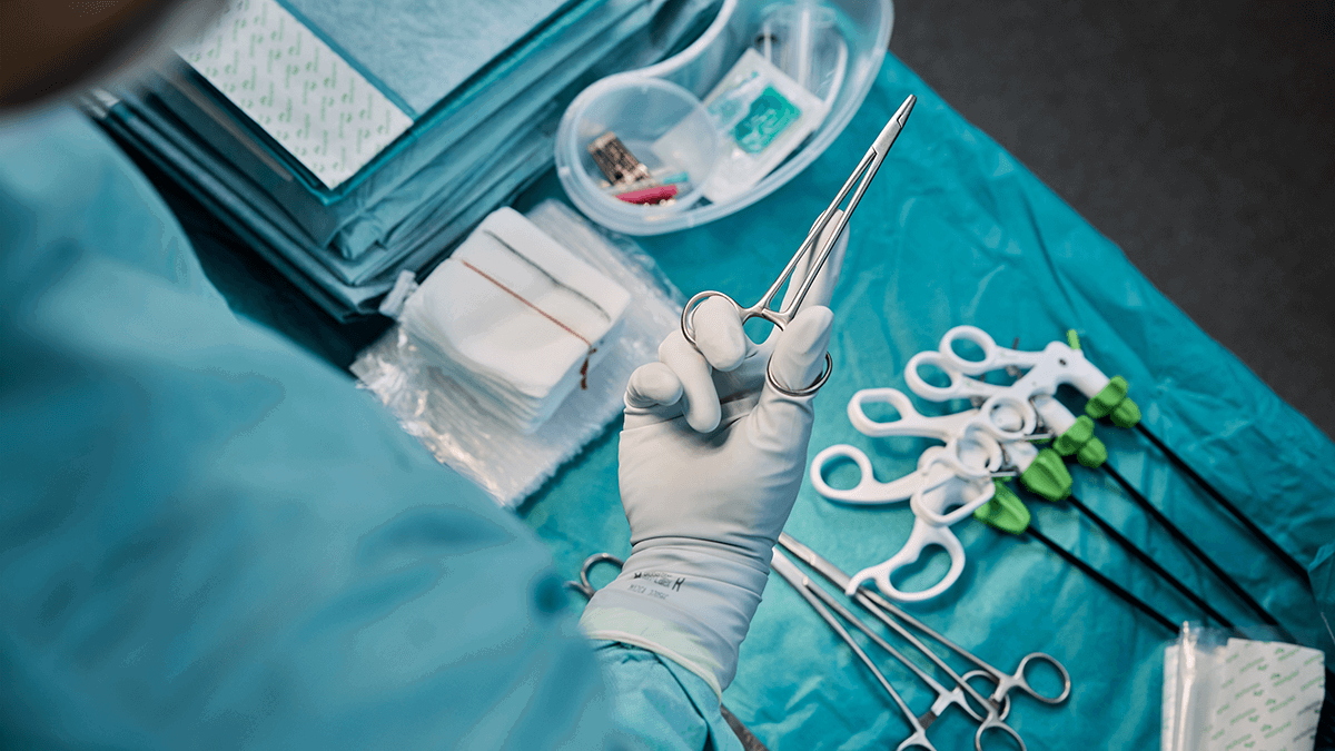 The hand of a surgeon holding a clamp over other surgical tools and draping laid out on a table. Photo. 