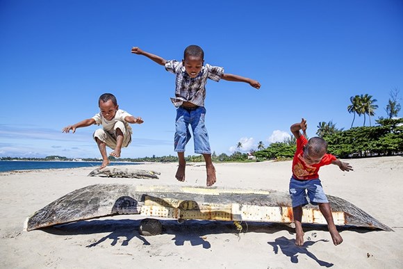 three boys jumping over a boat by a sea shore smiling